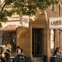 Caf&eacute; Lang an einer Stra&szlig;enecke mit Menschen, die drau&szlig;en sitzen. Ein 30er-Zone-Schild und B&auml;ume sind ebenfalls zu sehen., &copy; SMG, Sarah Schmid