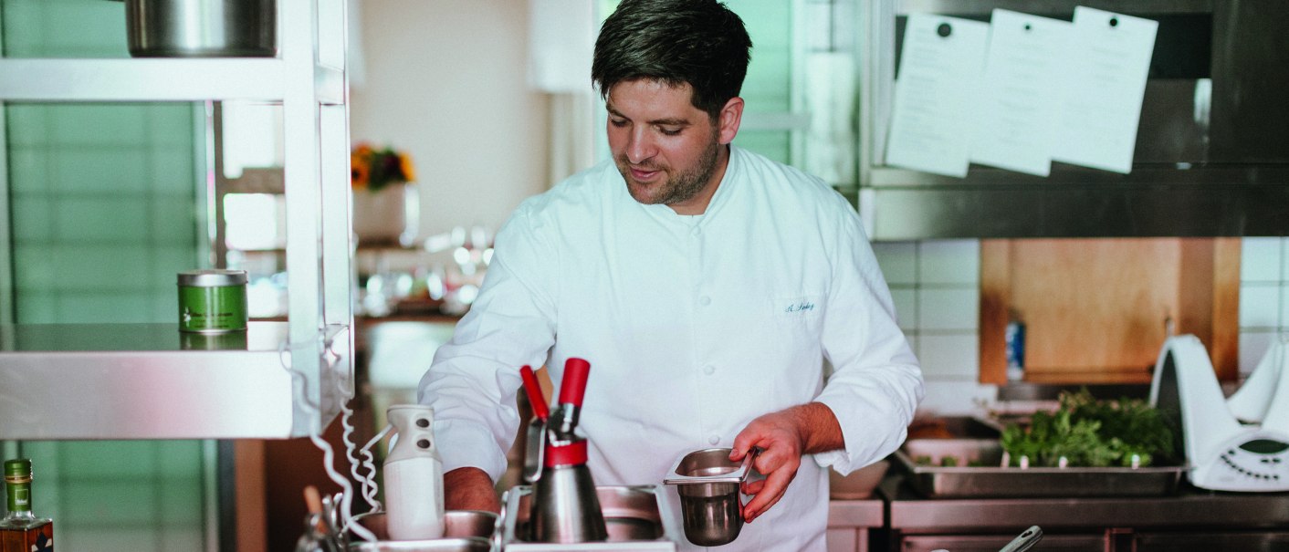 Ein Koch in weißer Uniform arbeitet konzentriert in einer professionellen Küche mit Töpfen und Utensilien., © Daniela Reske