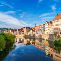 Fachwerkhäuser und moderne Gebäude spiegeln sich im Fluss in Schwäbisch Hall. Klare blaue Himmel und grüne Vegetation rahmen die Szene ein., © Michael Kühneisen