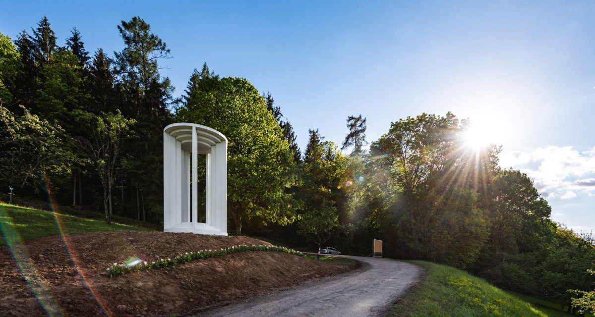 Weiße Säulenstruktur auf einem Hügel neben einem Weg, umgeben von Bäumen und Sonnenschein., © Authentic Studios Weiße Säulenstruktur auf einem Hügel neben einem Weg, umgeben von Bäumen und Sonnenschein., © Authentic Studios