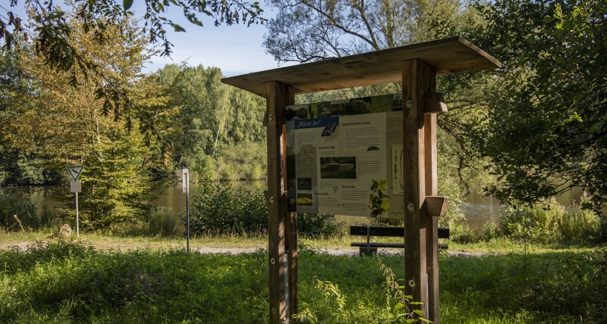 Infotafel am HölzerSee in einem bewaldeten Gebiet. Im Hintergrund sind Bäume und ein See zu sehen. Die Tafel steht auf einer Wiese., © Natur.Nah. Schönbuch & Heckengäu Infotafel am HölzerSee in einem bewaldeten Gebiet. Im Hintergrund sind Bäume und ein See zu sehen. Die Tafel steht auf einer Wiese., © Natur.Nah. Schönbuch & Heckengäu
