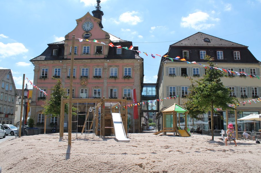 Ein Spielplatz mit Rutsche und Kletterger&uuml;st auf einem sandigen Platz vor historischen Geb&auml;uden, geschm&uuml;ckt mit bunten Wimpeln unter blauem Himmel., &copy; F&uuml;r die Bildrechte: Amt f&uuml;r Medien und Kommunikation Schw&auml;bisch Gm&uuml;nd