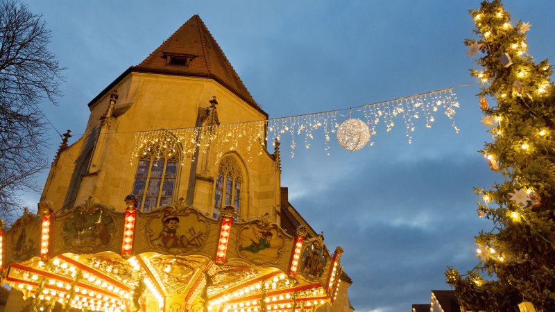 Eine festlich beleuchtete Stadtkirche mit Lichterketten, einem Karussell und einem geschmückten Weihnachtsbaum im Vordergrund., © Stadt Nürtingen