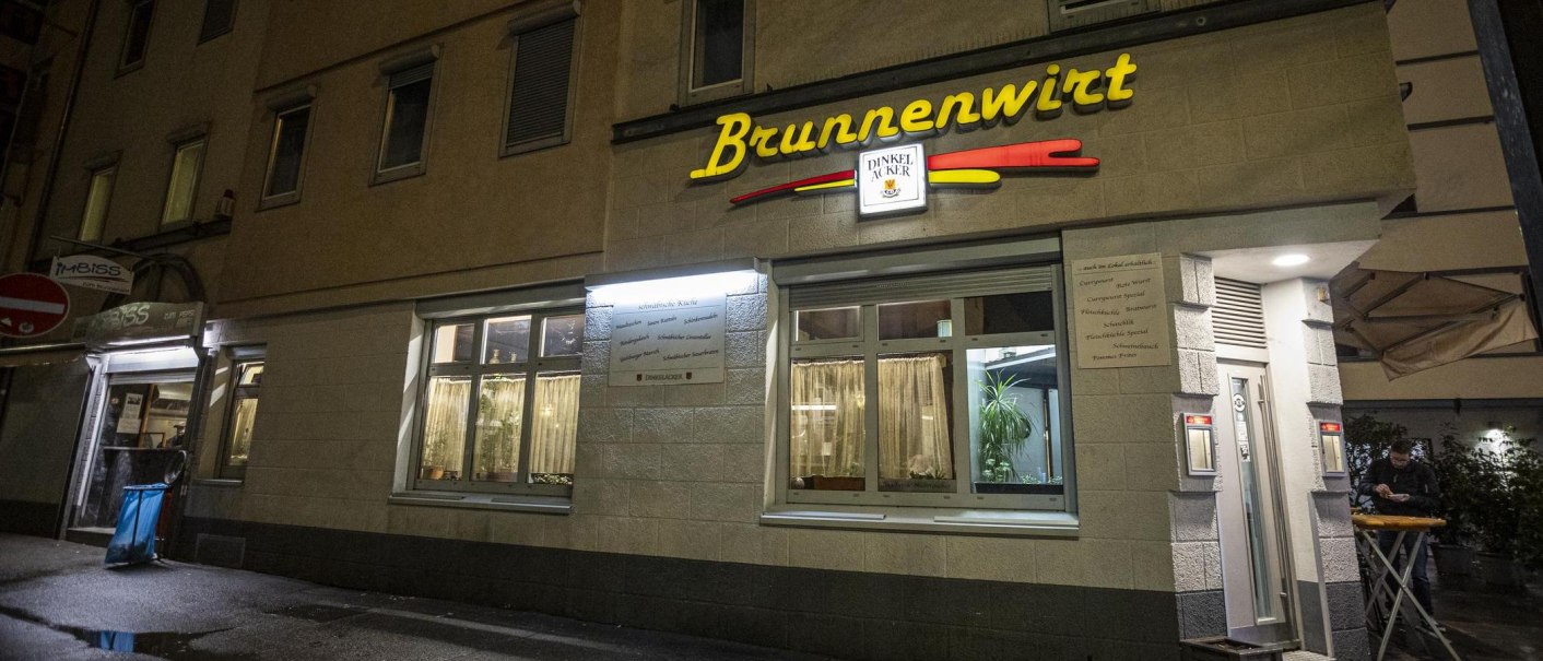 Das Restaurant Brunnenwirt bei Nacht, beleuchtet mit gelbem Schriftzug. Ein Mann steht an einem Tisch vor dem Eingang., © SMG, Sarah Schmid Das Restaurant Brunnenwirt bei Nacht, beleuchtet mit gelbem Schriftzug. Ein Mann steht an einem Tisch vor dem Eingang., © SMG, Sarah Schmid