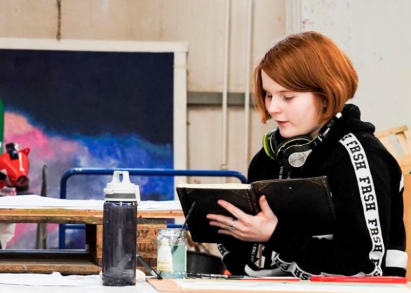 Eine Person mit Kopfhörern liest in einem Notizbuch. Im Hintergrund sind Malutensilien und ein farbenfrohes Bild zu sehen., © Kunstschule Labyrinth