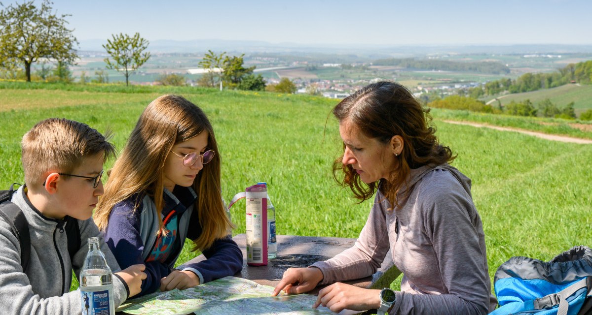 Drei Personen sitzen an einem Tisch im Freien und betrachten eine Karte. Im Hintergrund ist eine weite Landschaft mit Feldern und Bäumen zu sehen., © Stadtverwaltung Winnenden