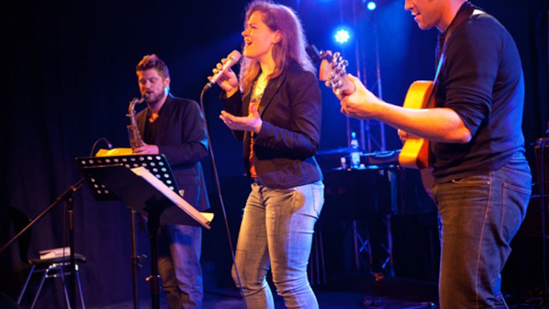 Drei Musiker auf der B&uuml;hne: Eine Frau singt, begleitet von einem Gitarristen und einem Saxophonisten. Die B&uuml;hne ist in blaues Licht getaucht., &copy; Jazzclub Schw&auml;bisch Hall e.V.