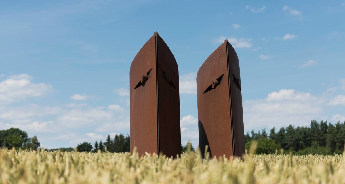 Zwei rostige, abstrakte Skulpturen stehen in einem Feld unter blauem Himmel. Im Hintergrund sind Bäume zu sehen., © Natur.Nah. Schönbuch & Heckengäu Zwei rostige, abstrakte Skulpturen stehen in einem Feld unter blauem Himmel. Im Hintergrund sind Bäume zu sehen., © Natur.Nah. Schönbuch & Heckengäu
