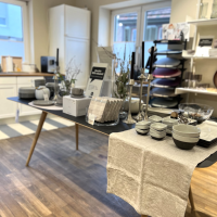 Ein Tisch in einem Laden mit Geschirr, Schalen, Kerzenhaltern und Vasen. Der Raum hat Holzboden und weiße Wände mit Regalen., © schöner laden Ein Tisch in einem Laden mit Geschirr, Schalen, Kerzenhaltern und Vasen. Der Raum hat Holzboden und weiße Wände mit Regalen., © schöner laden