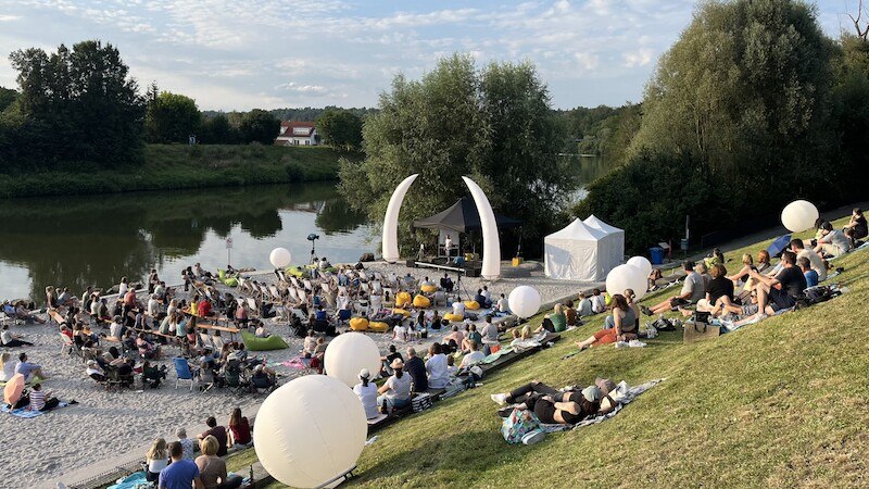 Open-Air-Poetry Slam am Flussufer mit vielen Zuschauern auf einer Wiese und einem Strandbereich. Eine B&uuml;hne mit dekorativen B&ouml;gen ist aufgebaut., &copy; Stadt Remseck am Neckar