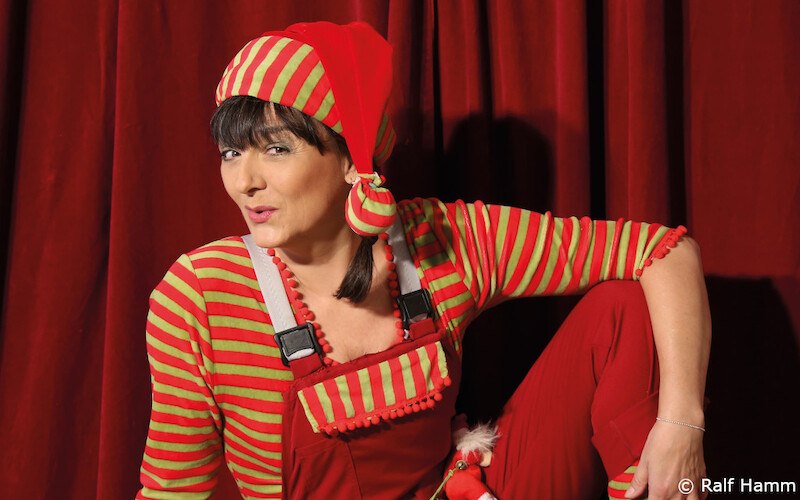 Eine Person in einem rot-grün gestreiften Outfit mit Mütze posiert vor einem roten Vorhang., © Kultur- und Sportamt Stadt Bietigheim-Bissingen
