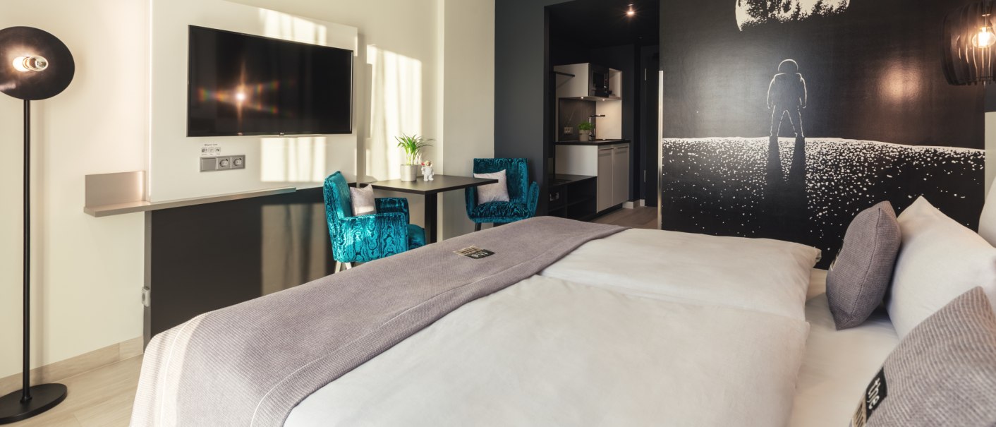 Modernes Zimmer mit großem Bett, blauen Sesseln, Astronauten-Wandbild, Flachbildfernseher und kleiner Küche., © Novum Management GmbH Modernes Zimmer mit großem Bett, blauen Sesseln, Astronauten-Wandbild, Flachbildfernseher und kleiner Küche., © Novum Management GmbH