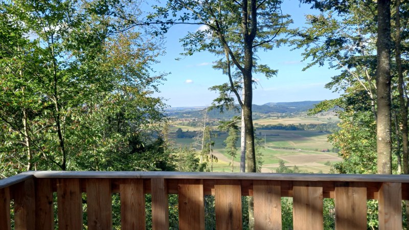 Holzgeländer mit Blick auf eine weite, grüne Landschaft. Felder und Hügel erstrecken sich bis zum Horizont, umgeben von Bäumen., © Petra Natzkowski Holzgeländer mit Blick auf eine weite, grüne Landschaft. Felder und Hügel erstrecken sich bis zum Horizont, umgeben von Bäumen., © Petra Natzkowski