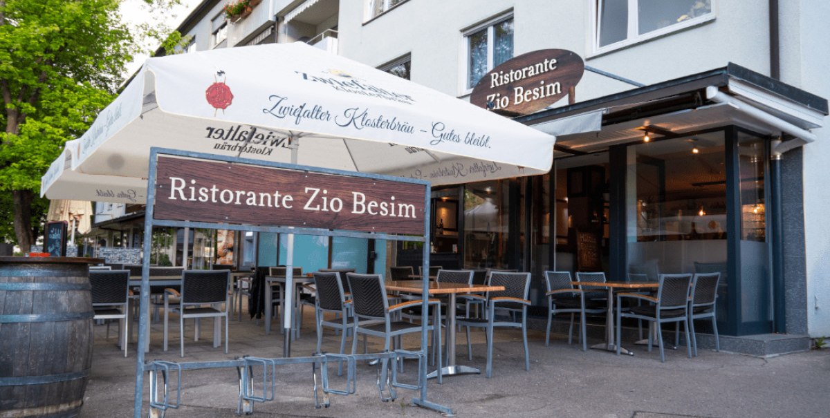 Außenbereich des Ristorante Zio Besim mit Tischen, Stühlen und einem großen Sonnenschirm. Das Restaurant hat eine moderne Glasfassade.