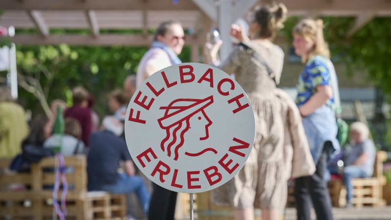 Ein Schild mit der Aufschrift 'Fellbach Erleben' steht im Vordergrund. Im Hintergrund sind Menschen bei einer Outdoor-Veranstaltung zu sehen., &copy; Stadt Fellbach