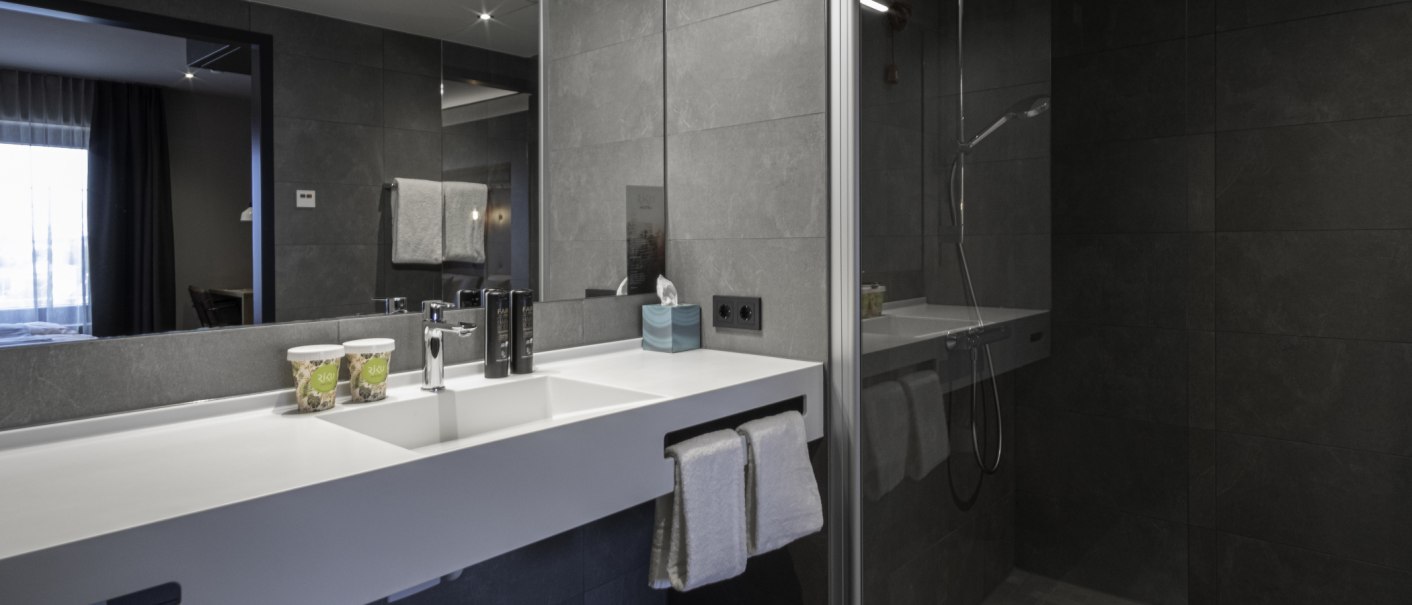 Modernes Badezimmer mit grauen Fliesen, großem Spiegel, Waschbecken und Dusche. Handtücher und Pflegeprodukte sind vorhanden., © 13 6 Design GmbH (Judenhof 10, 89073 Ulm)