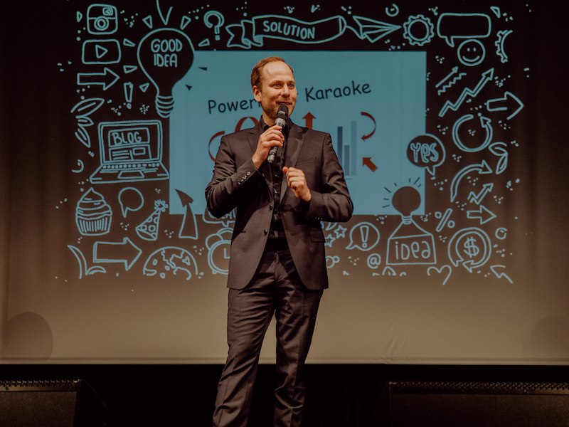 Ein Mann im Anzug spricht auf einer Bühne vor einer PowerPoint-Folie mit dem Titel 'PowerPoint Karaoke' und verschiedenen Symbolen., © Stadt Leinfelden-Echterdingen, Kulturamt
