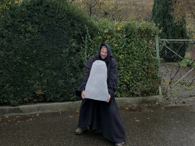 Ein Mann in einem dunklen Kost&uuml;m h&auml;lt einen gro&szlig;en, grauen Stein. Er steht auf einer nassen Stra&szlig;e vor einer Hecke., &copy; Cool-Tours StattReisen