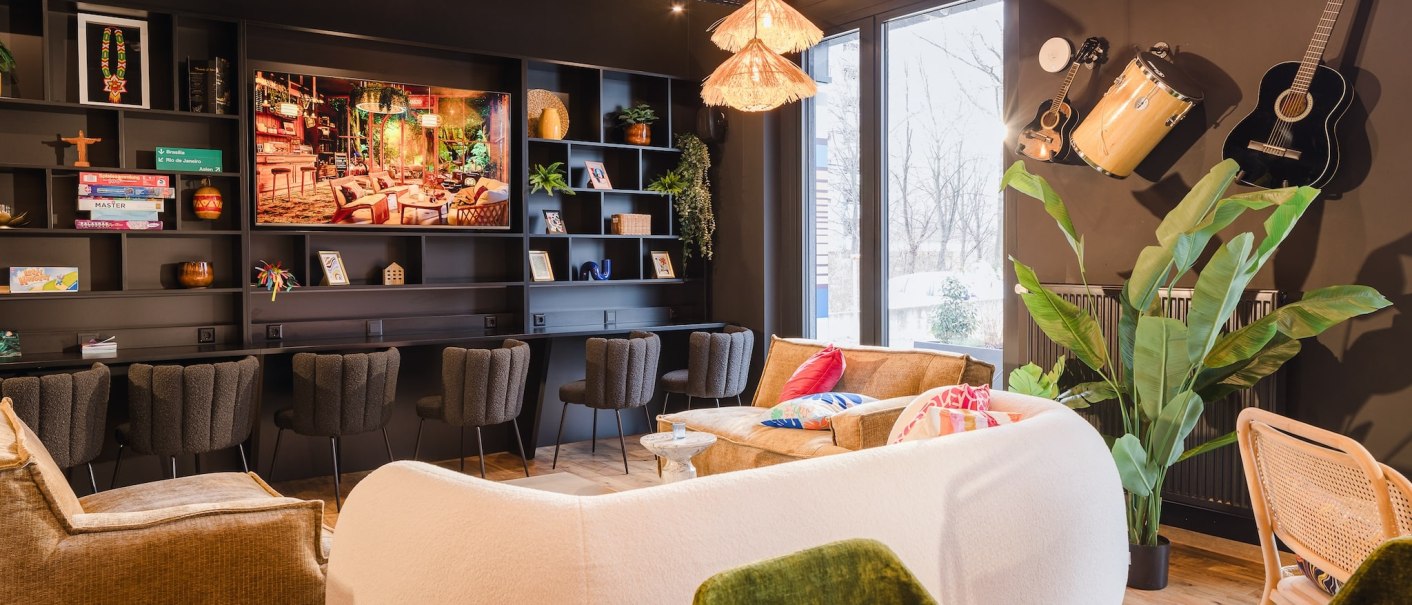 Ein gemütlicher Raum mit Sofas, Regalen voller Bücher und Spiele, Musikinstrumenten an der Wand und großen Fenstern. Pflanzen und warme Beleuchtung schaffen eine einladende Atmosphäre., © TOMAS Ein gemütlicher Raum mit Sofas, Regalen voller Bücher und Spiele, Musikinstrumenten an der Wand und großen Fenstern. Pflanzen und warme Beleuchtung schaffen eine einladende Atmosphäre., © TOMAS