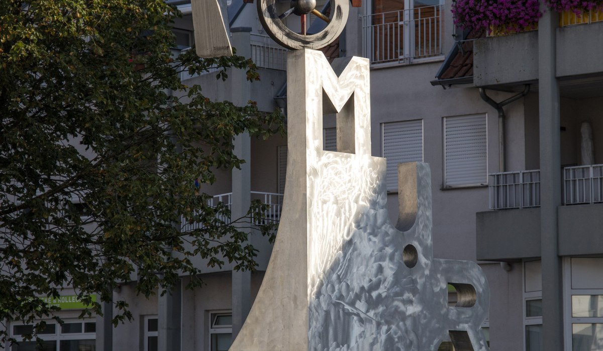 Der Katz- und Mausbrunnen in Bottwartal zeigt eine moderne Skulptur mit Buchstaben und geometrischen Formen vor einem Gebäude mit roten Dachziegeln., © Stuttgart Marketing