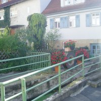 Haus mit blauen Fensterläden, umgeben von Pflanzen und Blumen. Ein grünes Geländer führt über einen kleinen Graben., © Natur.Nah. Schönbuch & Heckengäu Haus mit blauen Fensterläden, umgeben von Pflanzen und Blumen. Ein grünes Geländer führt über einen kleinen Graben., © Natur.Nah. Schönbuch & Heckengäu