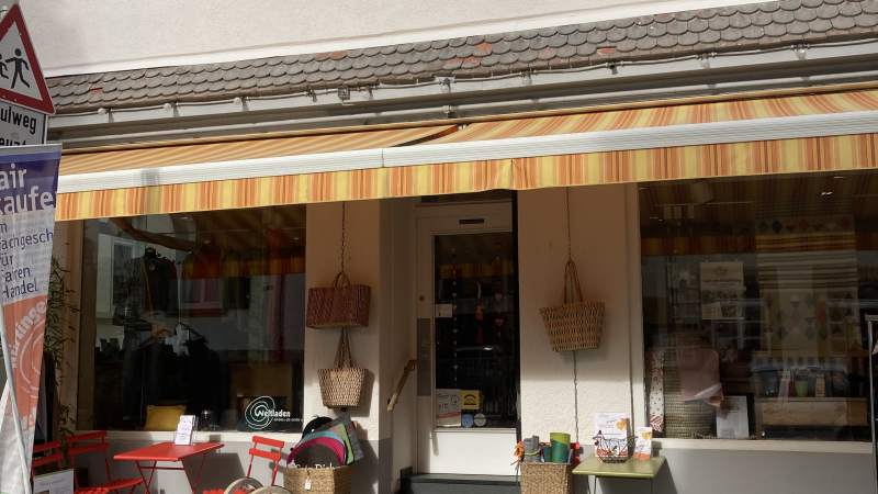 Ein kleiner Laden mit bunten Tischen und Stühlen vor der Tür. Eine Markise spendet Schatten. Im Schaufenster sind Körbe und Dekorationen zu sehen., © L. Besmer