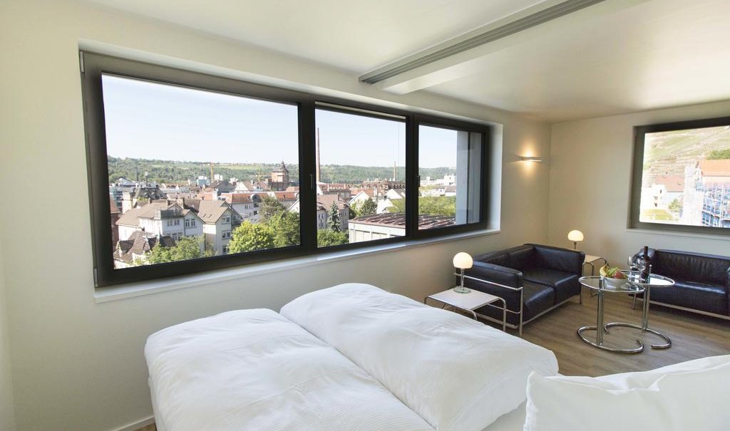 Modernes Apartment mit großem Fenster, Blick auf die Stadt, Doppelbett, schwarzem Sofa und kleinem Tisch mit Lampe., © Büroma-Apart Apartmentvermietung GmbH Modernes Apartment mit großem Fenster, Blick auf die Stadt, Doppelbett, schwarzem Sofa und kleinem Tisch mit Lampe., © Büroma-Apart Apartmentvermietung GmbH