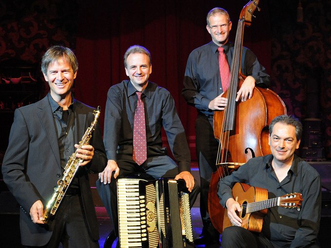 Eine Jazzband mit Saxophon, Akkordeon, Kontrabass und Gitarre posiert auf einer Bühne. Die Musiker tragen dunkle Anzüge und lächeln in die Kamera., © Stadt Murrhardt