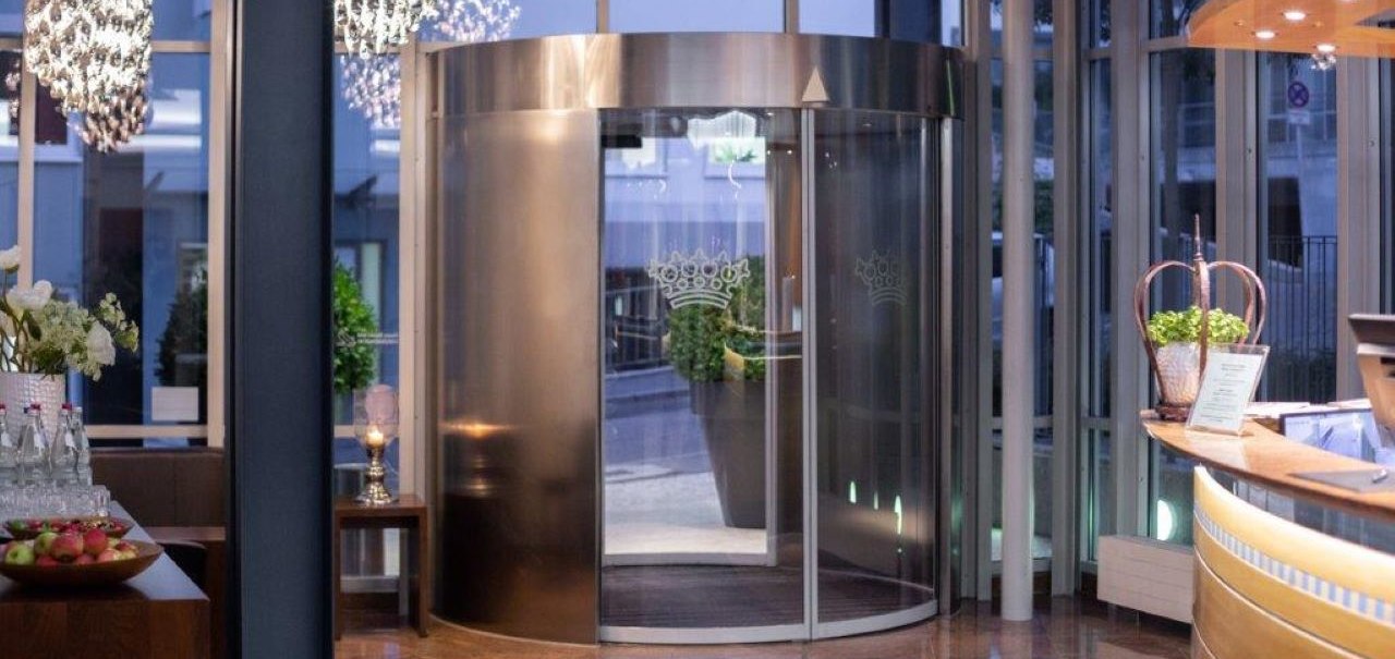 Moderner Eingangsbereich mit Drehtür, Empfangstresen und eleganter Beleuchtung. Dekorative Pflanzen und Getränke auf einem Tisch., © Kronen Hotel GmbH