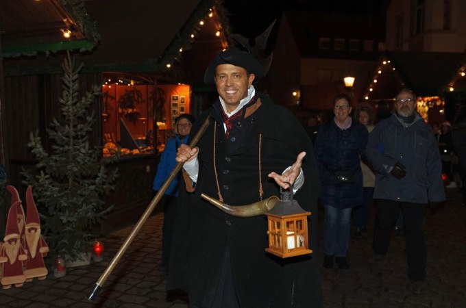 Ein Nachtwächter in traditioneller Kleidung mit Laterne und Horn führt eine Gruppe von Menschen über einen festlich beleuchteten Weihnachtsmarkt., © Stadt Nürtingen Ein Nachtwächter in traditioneller Kleidung mit Laterne und Horn führt eine Gruppe von Menschen über einen festlich beleuchteten Weihnachtsmarkt., © Stadt Nürtingen