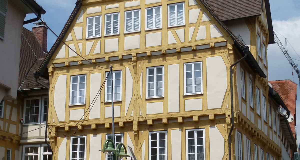 Ein historisches Fachwerkhaus mit gelben Balken, Uhr und Glockenturm. Es steht an einer gepflasterten Straße mit Parkscheinautomat und Blumenkübel. Ein historisches Fachwerkhaus mit gelben Balken, Uhr und Glockenturm. Es steht an einer gepflasterten Straße mit Parkscheinautomat und Blumenkübel.