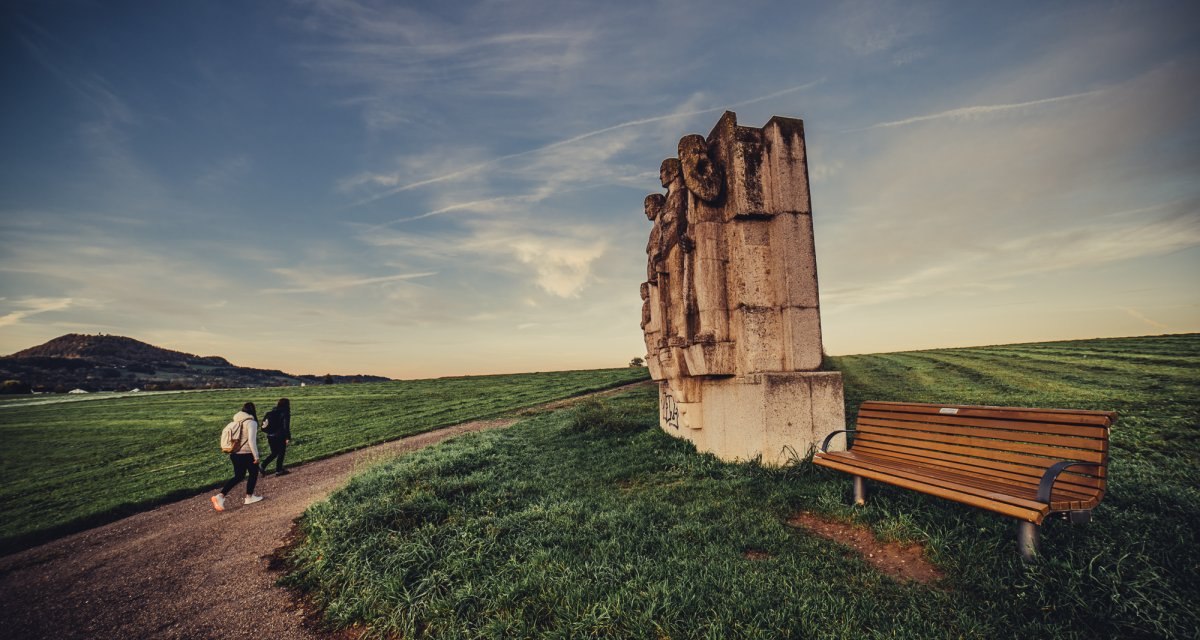 Zwei Personen spazieren auf einem Weg neben einer großen Steinskulptur und einer Bank auf einem grünen Feld unter blauem Himmel., © Landratsamt Ostalbkreis Zwei Personen spazieren auf einem Weg neben einer großen Steinskulptur und einer Bank auf einem grünen Feld unter blauem Himmel., © Landratsamt Ostalbkreis