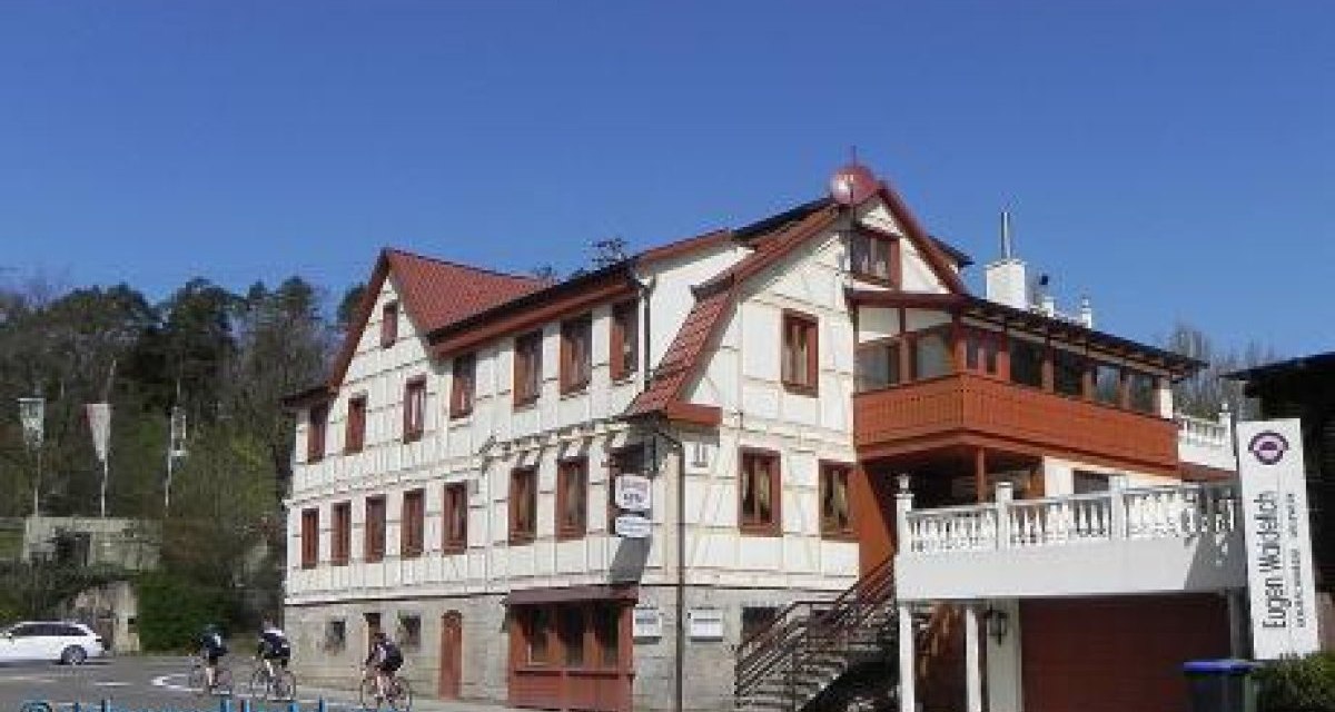 Ein traditionelles Fachwerkgebäude mit rotem Dach und Balkon. Drei Radfahrer fahren vorbei. Der Himmel ist klar und blau., © Natur.Nah. Schönbuch & Heckengäu Ein traditionelles Fachwerkgebäude mit rotem Dach und Balkon. Drei Radfahrer fahren vorbei. Der Himmel ist klar und blau., © Natur.Nah. Schönbuch & Heckengäu