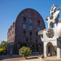 Moderne Skulptur mit Pferde- und Menschenköpfen vor einem Backsteingebäude mit rundem Dach, umgeben von Bäumen und Pflanzen., © Stuttgart-Marketing GmbH, Achim Mende