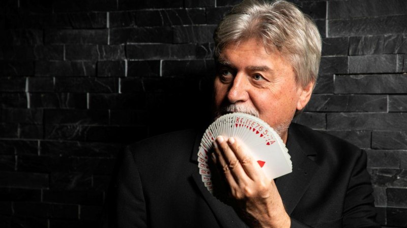 Mann mit grauen Haaren hält Spielkartenfächer vor Gesicht, dunkle Steinwand im Hintergrund., © Horst Reutter Mann mit grauen Haaren hält Spielkartenfächer vor Gesicht, dunkle Steinwand im Hintergrund., © Horst Reutter