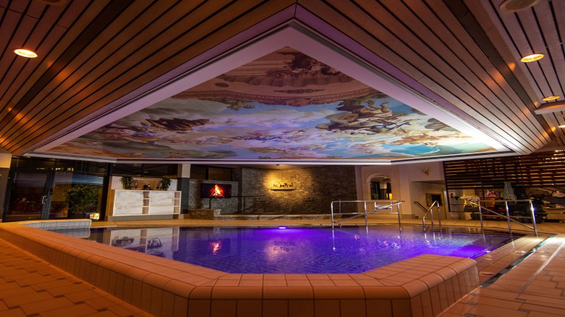 Ein beleuchtetes Hallenbad mit bemalter Decke und einem Pool in der Mitte. Die Decke zeigt ein kunstvolles Fresko., © AlbThermen Bad Urach Ein beleuchtetes Hallenbad mit bemalter Decke und einem Pool in der Mitte. Die Decke zeigt ein kunstvolles Fresko., © AlbThermen Bad Urach