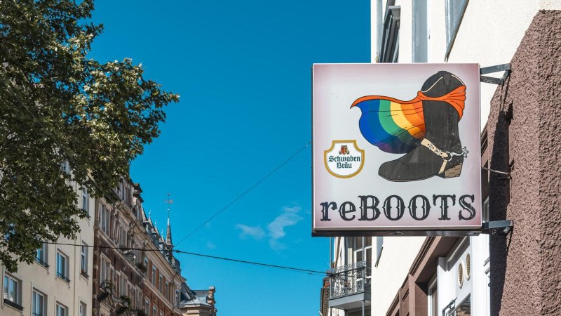 Ein Schild mit einem Stiefel und einer Regenbogenflagge, daneben die Aufschrift 'reBOOTS'. Im Hintergrund sind Gebäude und ein Baum zu sehen., © SMG, Sarah Schmid