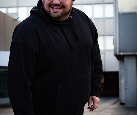 Ein Mann in schwarzem Hoodie und Mütze lächelt in einer städtischen Umgebung mit modernen Gebäuden im Hintergrund., © SBEntertainment GmbH & Co. KG