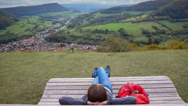 Person entspannt auf einer Holzbank mit Blick auf ein Tal, grüne Felder und ein Dorf. Ein roter Rucksack liegt daneben. Der Himmel ist bewölkt., © Foto: Mario Klaiber