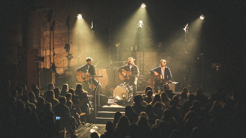 Drei Musiker spielen auf einer Bühne vor einem sitzenden Publikum. Die Bühne ist in warmes Licht getaucht, Instrumente und Mikrofone sind sichtbar., © Scala Kultur Live gGmbH