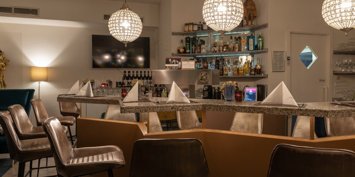 Elegante Bar mit beleuchtetem Tresen, Flaschenregal und modernen Hängelampen. Lederstühle und gefaltete Servietten auf dem Tresen., © Hotel Princess