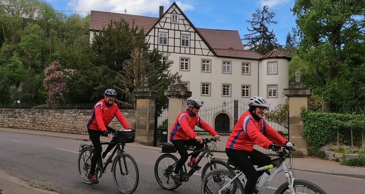 Drei Radfahrer in roten Jacken fahren auf einer Straße vor einem großen Fachwerkhaus mit Garten., © Land der 1000 Hügel - Kraichgau-Stromberg Drei Radfahrer in roten Jacken fahren auf einer Straße vor einem großen Fachwerkhaus mit Garten., © Land der 1000 Hügel - Kraichgau-Stromberg