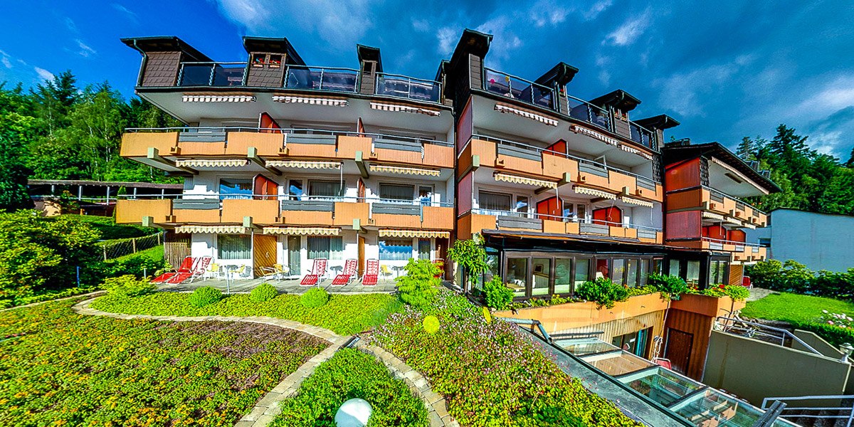 Mehrstöckiges Gebäude mit Balkonen, umgeben von gepflegtem Garten und blühenden Pflanzen unter blauem Himmel., © Hotel Hochwald