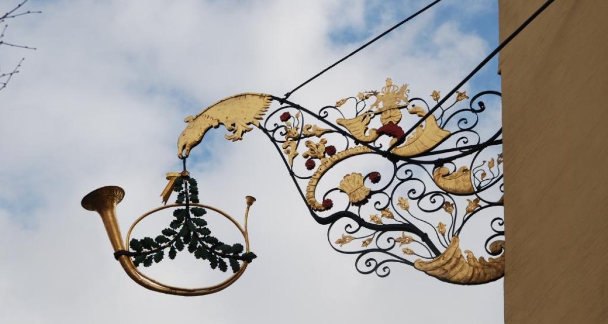 Ein kunstvoll verziertes Metallschild mit einem goldenen Waldhorn und dekorativen Elementen hängt an einer Hauswand vor blauem Himmel., © Ludwigsburg - Stuttgart-Marketing GmbH Ein kunstvoll verziertes Metallschild mit einem goldenen Waldhorn und dekorativen Elementen hängt an einer Hauswand vor blauem Himmel., © Ludwigsburg - Stuttgart-Marketing GmbH