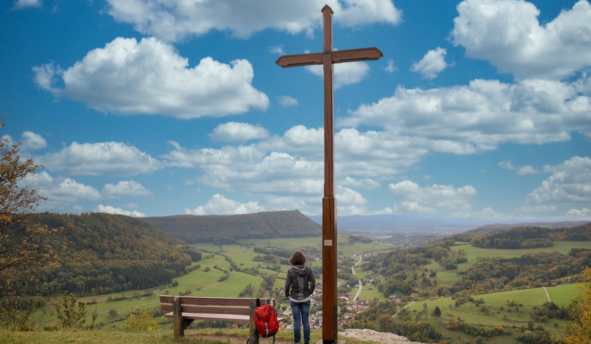 Person am Aussichtspunkt Kreuz Galgenberg, neben einem großen Holzkreuz, mit Blick auf eine weite grüne Landschaft und einen bewölkten blauen Himmel., © Foto: Mario Klaiber