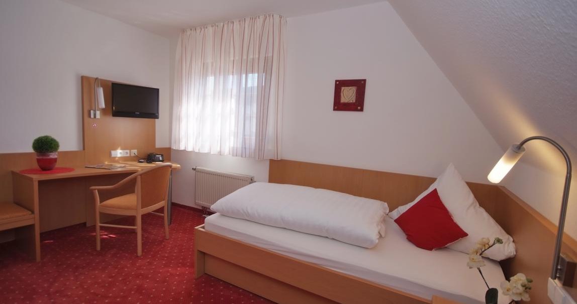 Kleines Hotelzimmer mit Einzelbett, rotem Teppich, Schreibtisch, Stuhl und Fernseher. Weiße Vorhänge und rote Akzente schaffen eine gemütliche Atmosphäre., © TOMAS