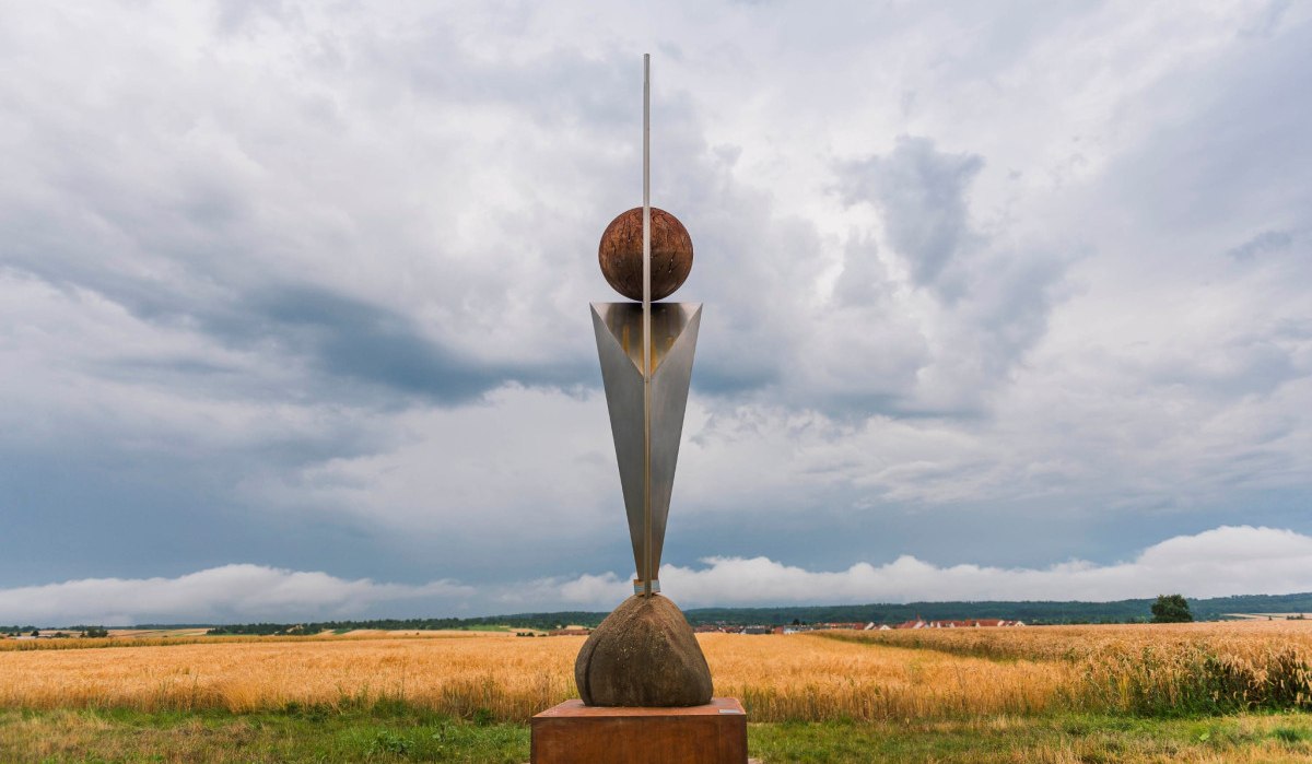 Eine moderne Skulptur steht auf einem Feld mit bewölktem Himmel im Hintergrund. Die Skulptur besteht aus geometrischen Formen und Metall., © Natur.Nah. Schönbuch & Heckengäu Eine moderne Skulptur steht auf einem Feld mit bewölktem Himmel im Hintergrund. Die Skulptur besteht aus geometrischen Formen und Metall., © Natur.Nah. Schönbuch & Heckengäu