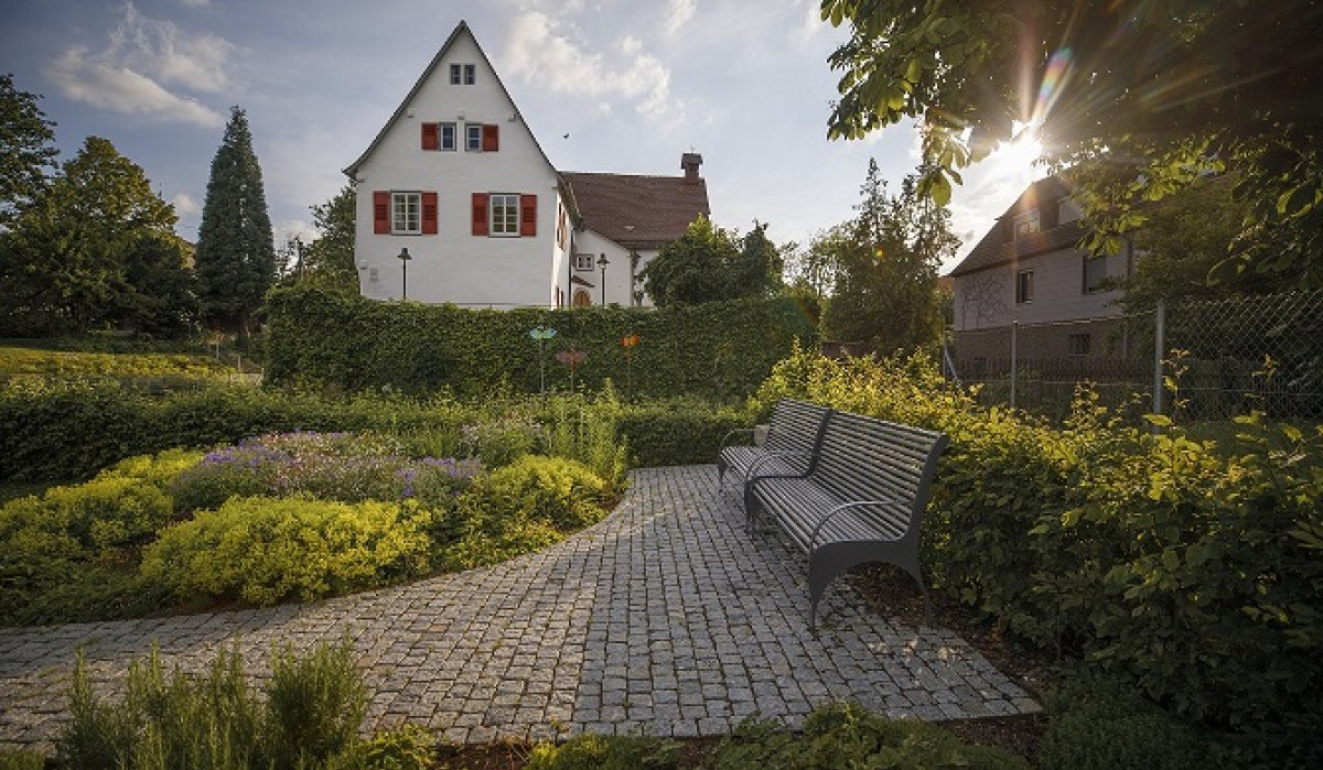 Ein gepflegter Garten mit Bänken, gepflastertem Weg und einem weißen Haus im Hintergrund. Die Sonne scheint durch die Bäume., © Stoppel Ein gepflegter Garten mit Bänken, gepflastertem Weg und einem weißen Haus im Hintergrund. Die Sonne scheint durch die Bäume., © Stoppel