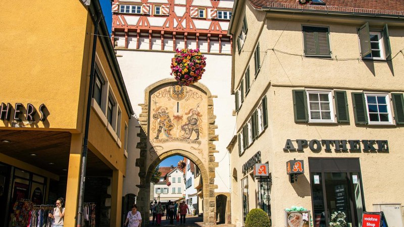 Das Stadttor von Bietigheim-Bissingen mit Fachwerk und Wandmalerei, umgeben von Geschäften wie einer Apotheke und einem Bekleidungsgeschäft., © Stuttgart-Marketing GmbH, Sarah Schmid Das Stadttor von Bietigheim-Bissingen mit Fachwerk und Wandmalerei, umgeben von Geschäften wie einer Apotheke und einem Bekleidungsgeschäft., © Stuttgart-Marketing GmbH, Sarah Schmid