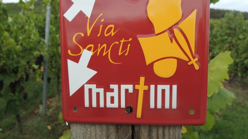 Rotes Schild mit 'Via Sancti Martini' und Pfeilen, befestigt an einem Holzpfosten in einem Weinberg., © Land der 1000 Hügel - Kraichgau-Stromberg